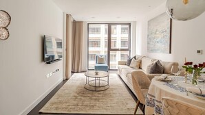 A modern living room with a sofa, a dining table set, a TV mounted on the wall, and a large window with curtains. - Luxury Riverfront Retreat in London (London)