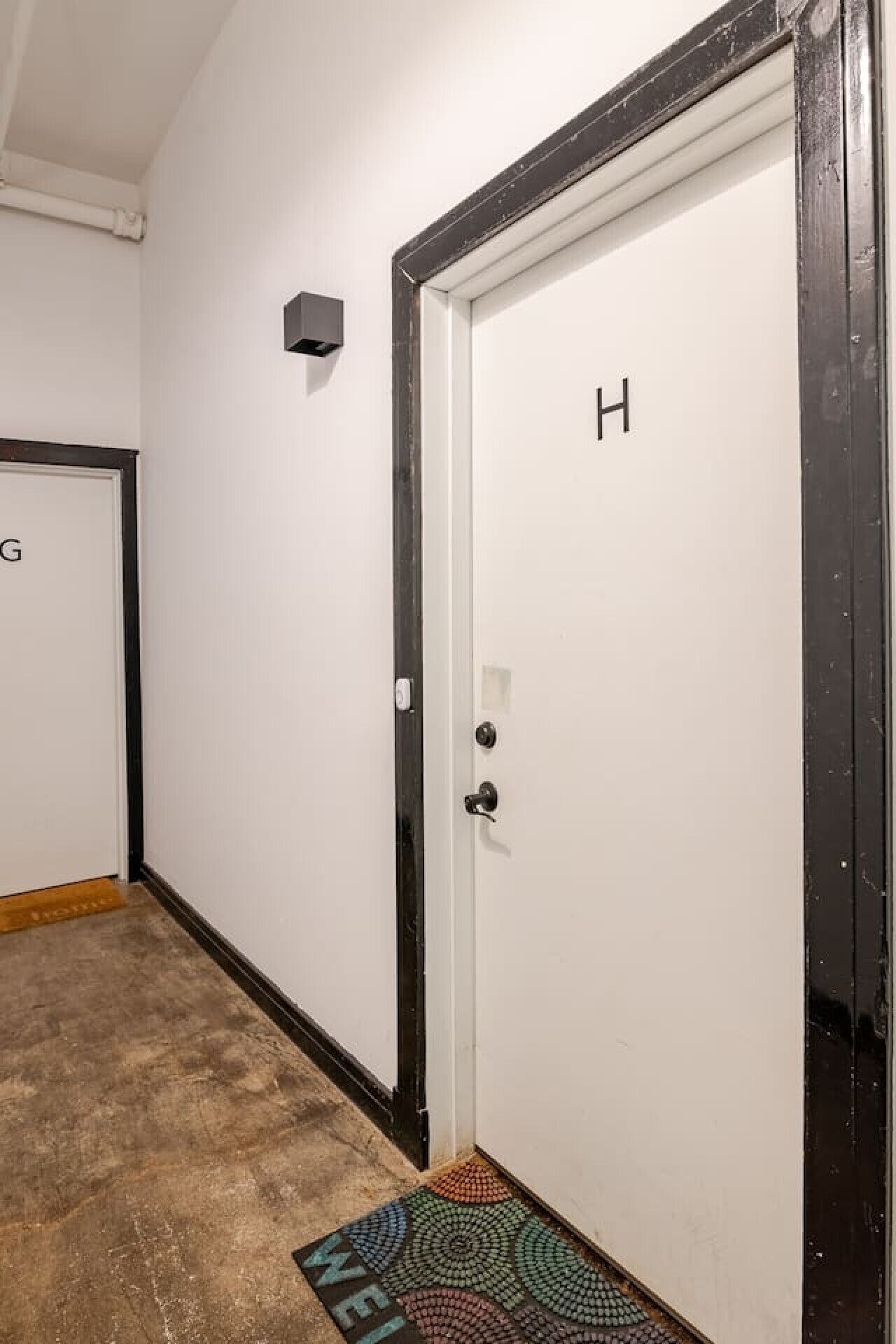 Een gang met een witte deur waarop de letter 'H' staat, een klein wandlampje en een kleurrijke deurmat.