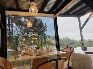 Un comedor con una mesa dispuesta para cuatro personas, rodeado de sillas de mimbre, bajo una estructura de vidrio con vistas a árboles.