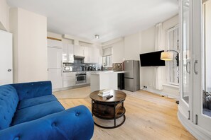 A modern living room with a blue sofa, a round coffee table, and a kitchen in the background. - Prime Soho 2BR/2BA Apartment with A/C (London)