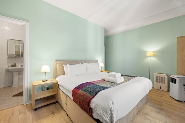 A hotel room with a bed, bedside tables, a nightstand, a lamp, a heater, and a bathroom with a mirror and sink.