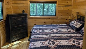 Ein Schlafzimmer in einer Holzhütte mit Holzbalkendecke, Deckenventilator, einem Bett mit gestecktem Quilz, einem Schminktisch und einem Fenster mit Blick auf einen Wald.