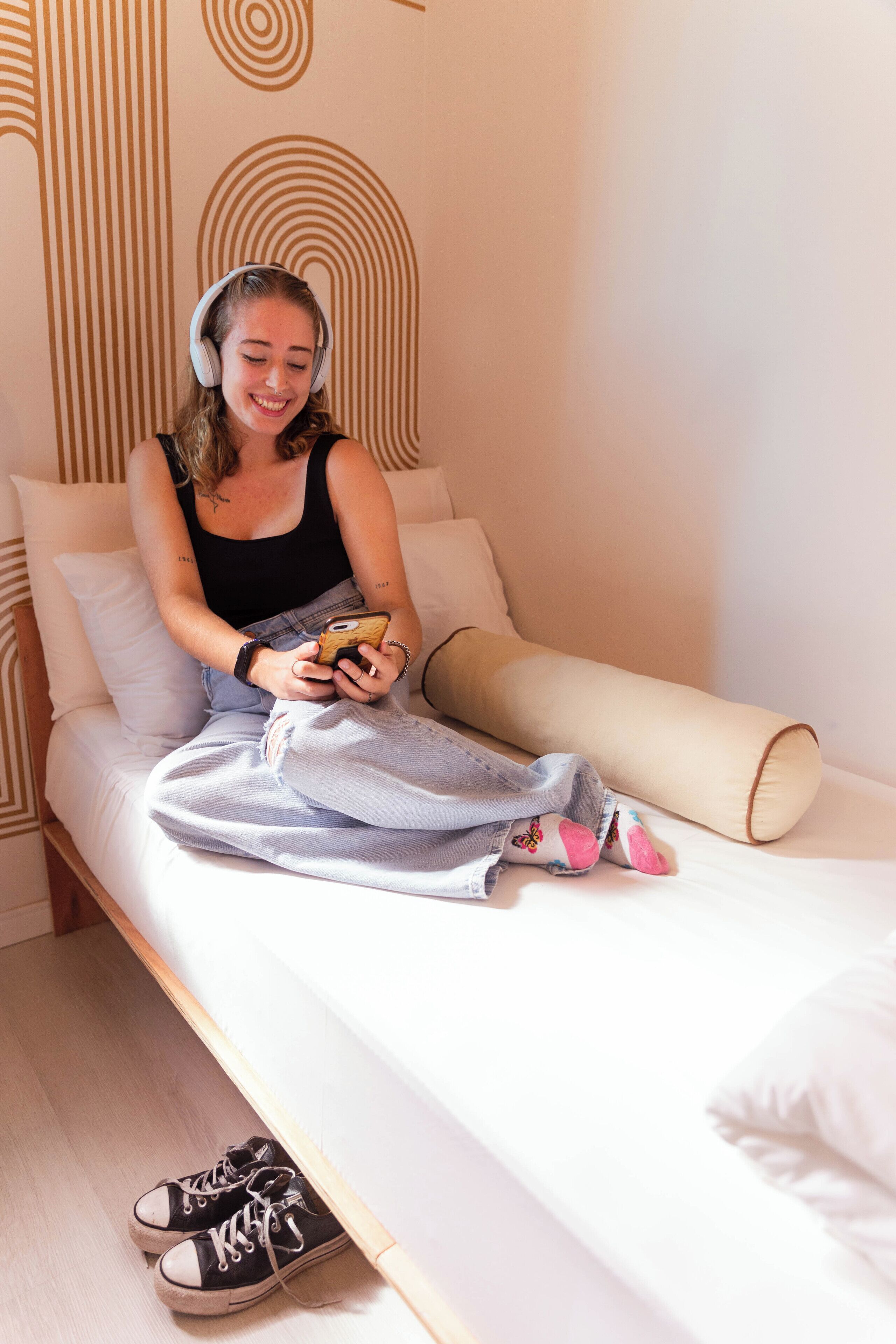 A person sitting on a bed with a smartphone, wearing headphones, and jeans.