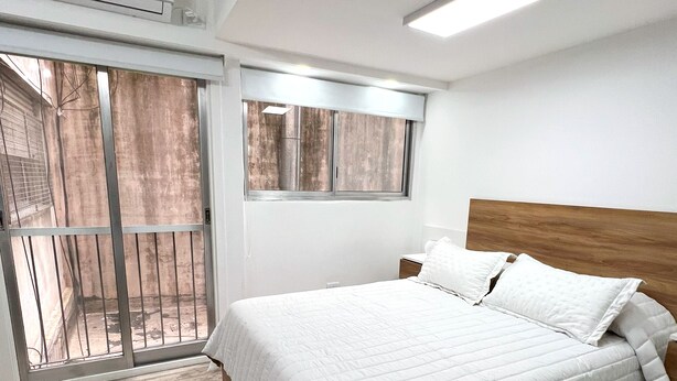 A bedroom with a wooden bed, white bedding, a large window, and an air conditioning unit.