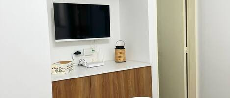 Una habitación moderna con un armario de madera, un televisor montado, una mesita con una cesta y una puerta al fondo.