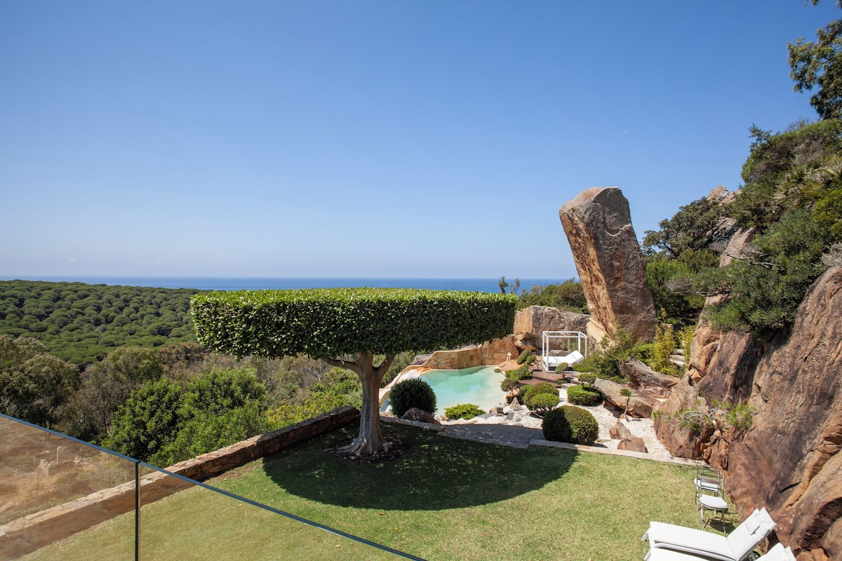 Un jardín ajardinado con piscina, una gran formación rocosa y vista al océano.