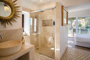 Un baño con un espejo con diseño de sol, un lavamanos de piedra, ducha con cerramiento de vidrio y vistas a un jardín a través de una puerta corrediza de cristal.