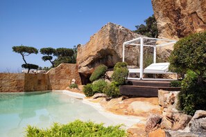 Un área de piscina con un telón de fondo de formación rocosa, una terraza de madera y una estructura de toldo blanco.