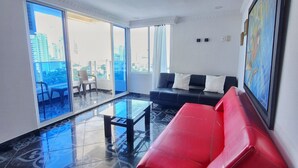 Ein modernes Wohnzimmer mit einem roten Sofa, einem Glastisch und einem schwarzen Schlafsofa. Große Fenster bieten Blick auf die Skyline der Stadt.