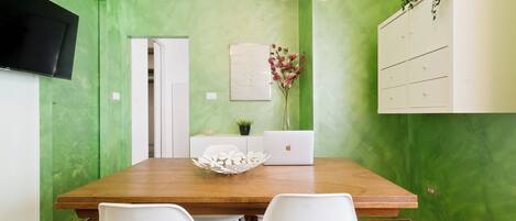 A dining area with a wooden table, white chairs, a laptop, a decorative bowl, a potted plant, and a wall-mounted TV.
