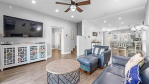 A modern living room with a flat-screen TV, a ceiling fan, a dining area, and a kitchen. - Purple Paradise (Pensacola)