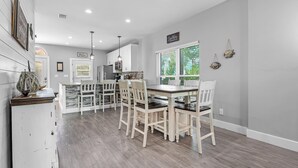 A modern kitchen with white cabinetry, a dining area with a wooden table and chairs, and a view of the outdoors through large windows. - Purple Paradise (Pensacola)