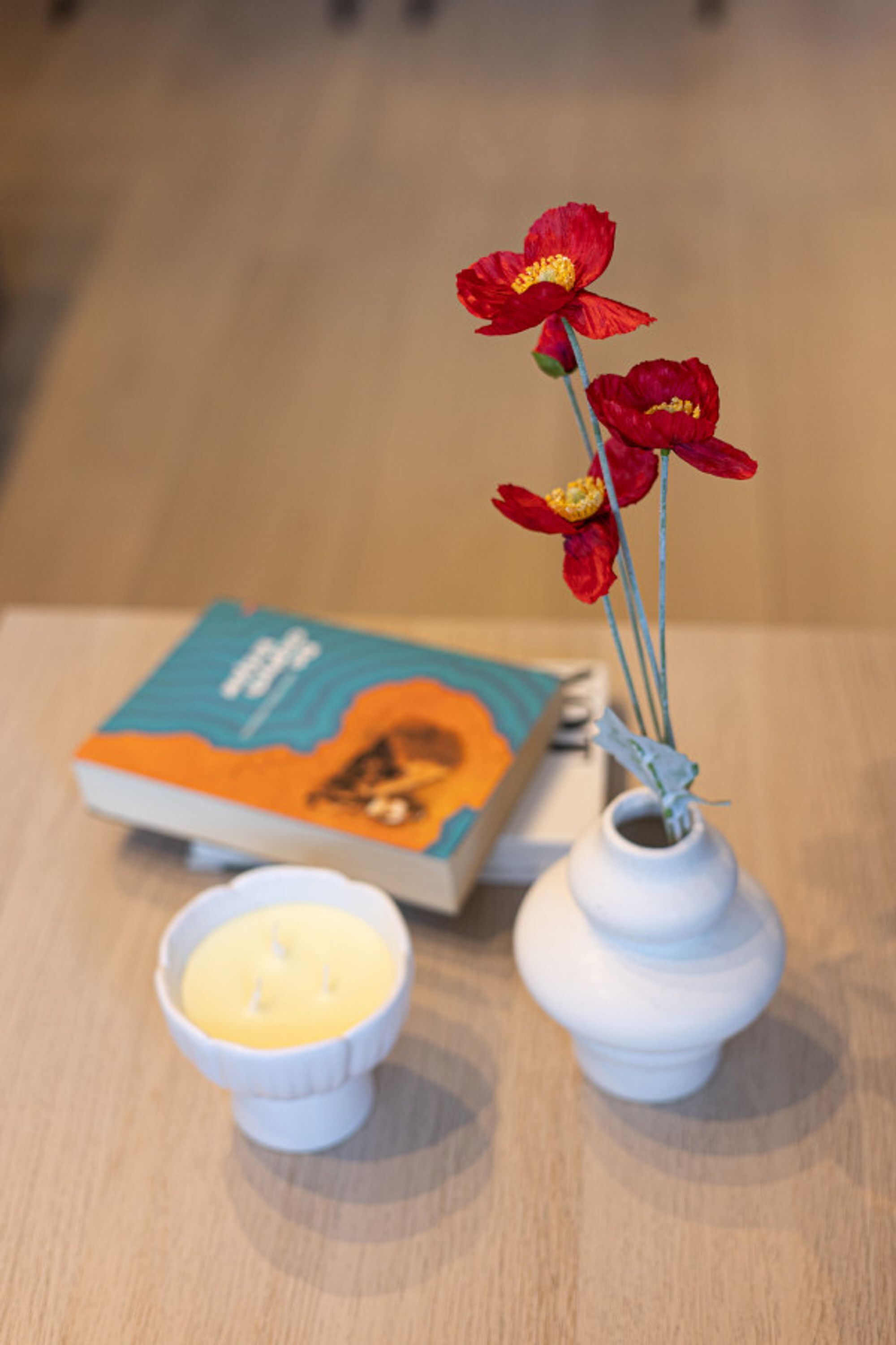 A white vase with red flowers and a book on a wooden table.