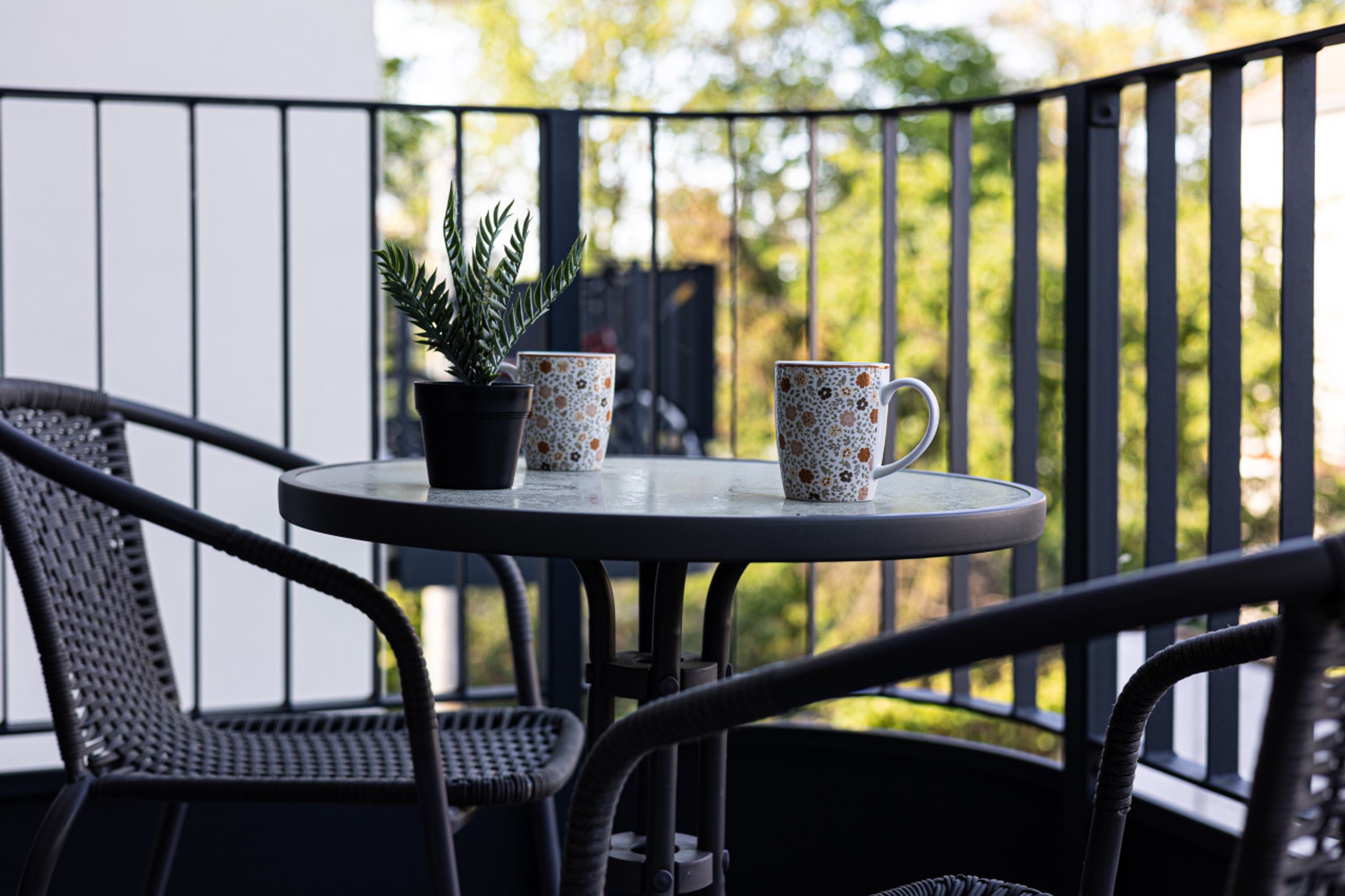 Une table ronde avec deux tasses décorées et une plante en pot, sur un balcon agrémenté de chaises en osier.