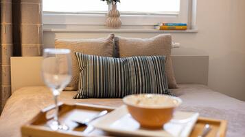 A neatly arranged breakfast tray with a glass of wine, a bowl of soup, and a plate of food on a bed with pillows and a vase of flowers by the window.