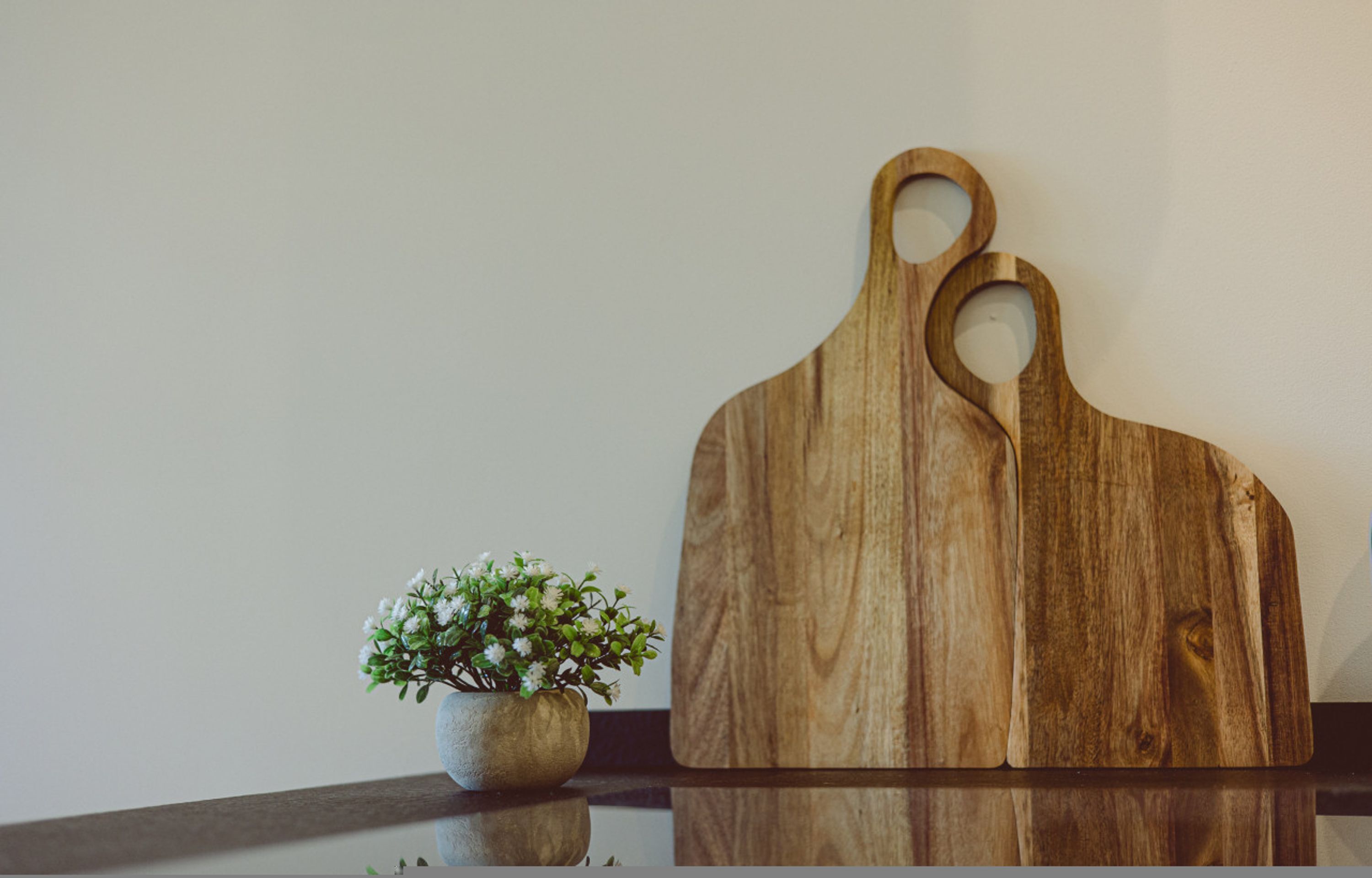 Ein Holzschneidebrett mit zwei Aussparungen, platziert auf einer spiegelnden Fläche neben einem kleinen Blumentopf mit weißen Blüten.