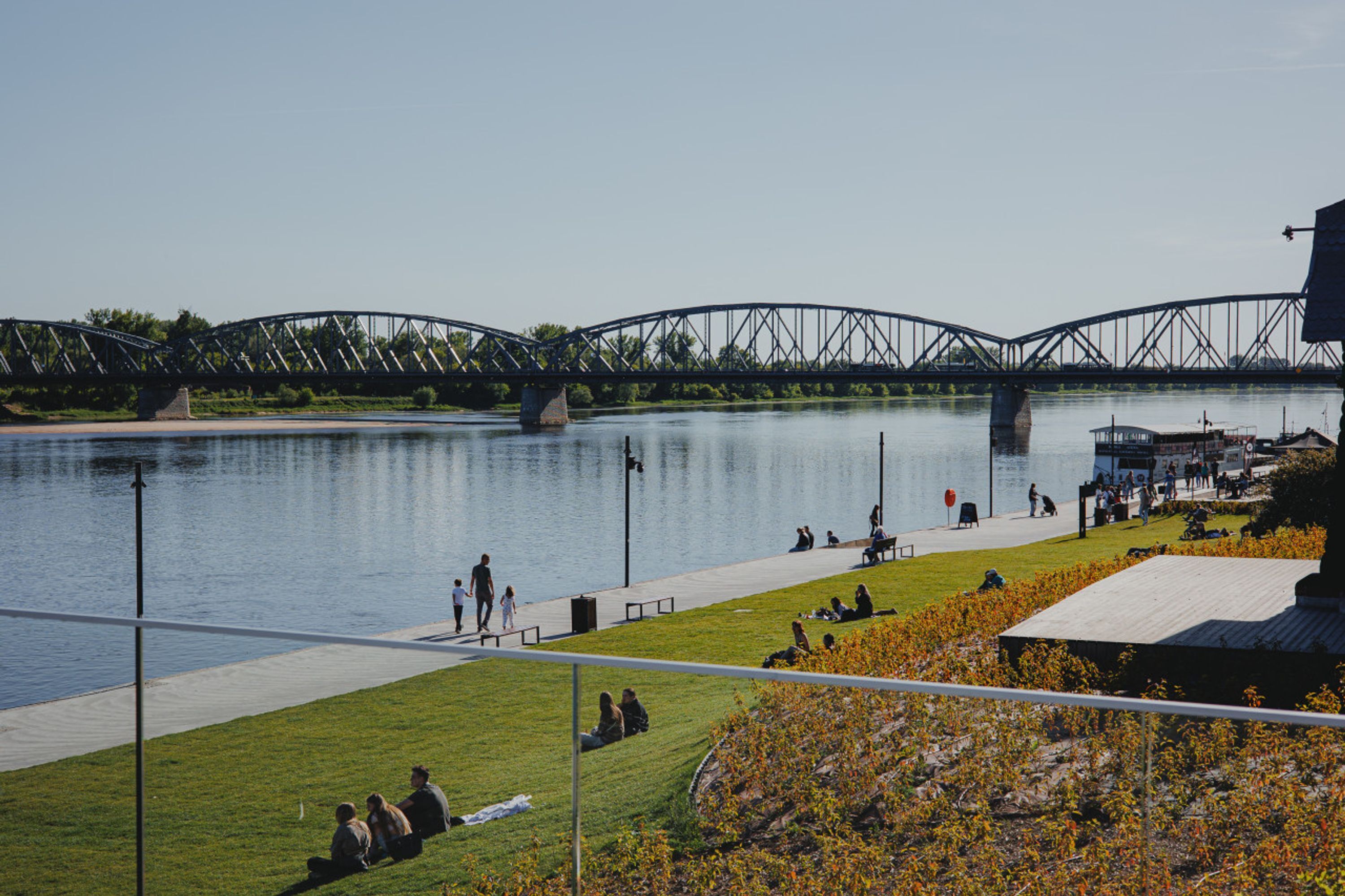 A riverside walkway with a bridge in the background, benches, and people relaxing.
