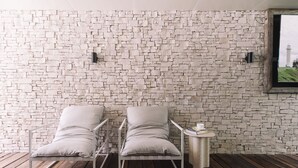 A room with a stone wall, two white metal chairs, a small table with a book and a cup, and a window with a view of a tower. - Bay Properties . Our Place (Yamba)