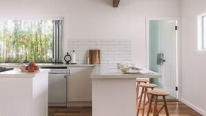A modern kitchen with white cabinetry, a wooden beam ceiling, a dishwasher, and a window with bamboo outside. - Bay Properties . Our Place (Yamba)
