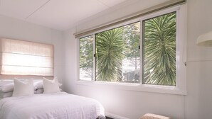 A bedroom with a large window, a bed, a bedside lamp, and a small ottoman. - Bay Properties . Our Place (Yamba)