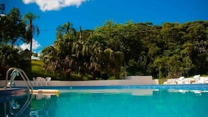 A clear blue swimming pool with a metal ladder, surrounded by white lounge chairs and lush greenery. - Recanto das Águas (Eldorado)