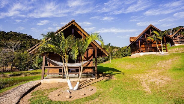 Two wooden cabins with thatched roofs, surrounded by lush greenery and a clear blue sky. - Recanto das Águas (Eldorado)