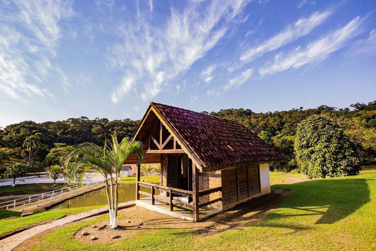 A wooden cabin with a red roof, surrounded by a pond and greenery.