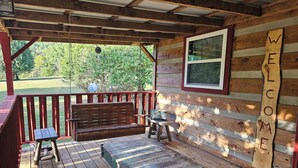 Una terraza de madera con un banco y una mesa, un cartel de bienvenida y una ventana con vistas a un paisaje verde.
