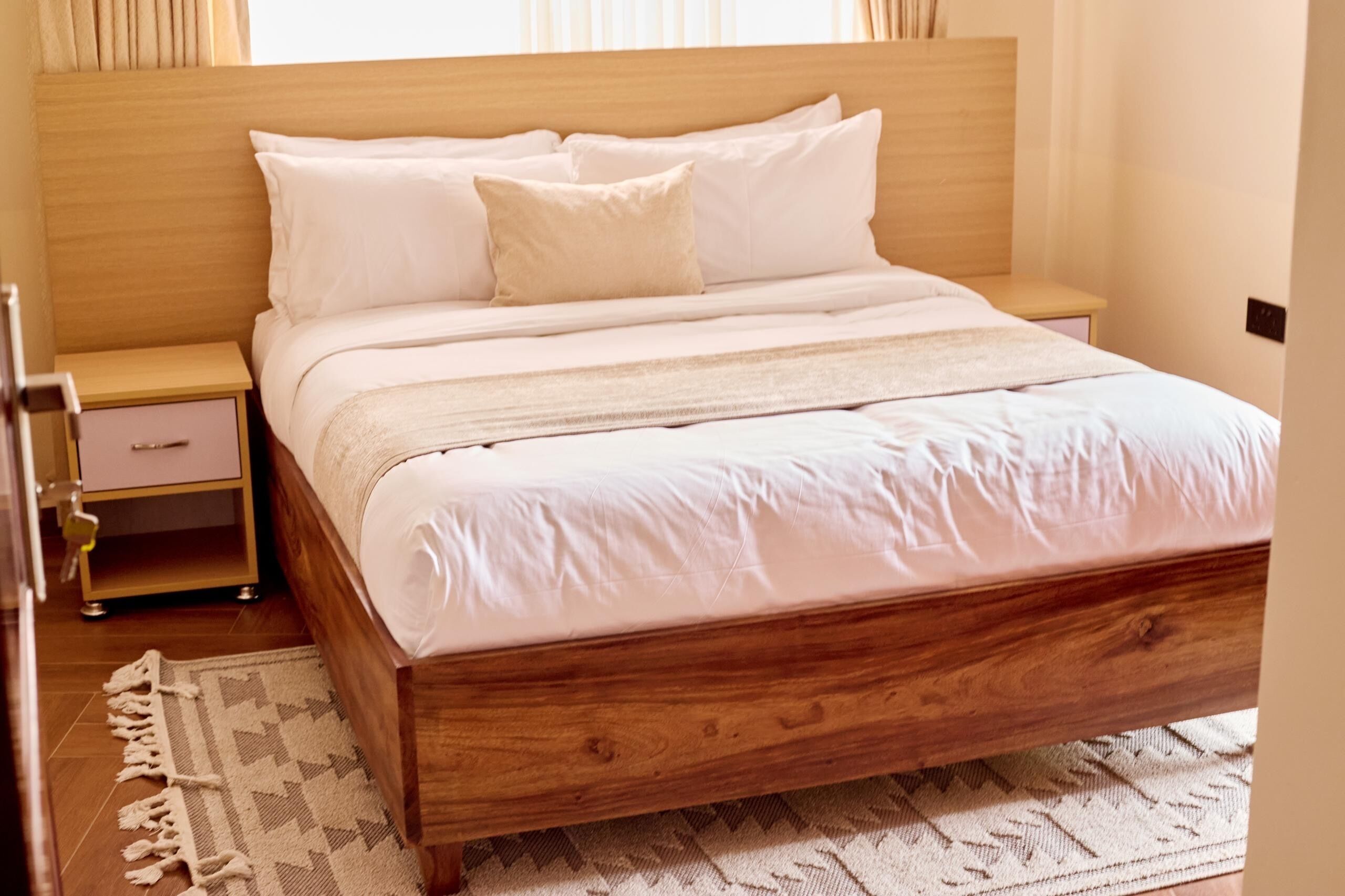 A neatly made bed with a wooden frame and white bedding, a bedside table with a lamp, and a patterned rug on the floor.
