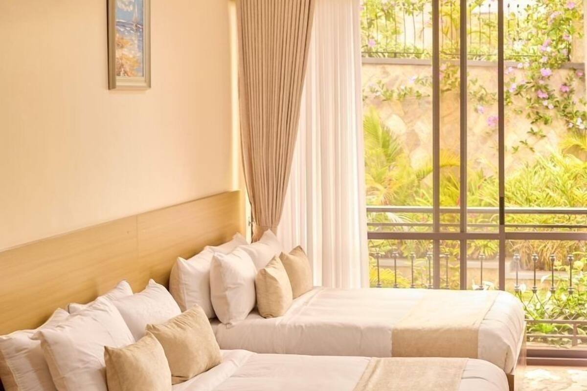 A hotel room with two beds, a large window with curtains, a wooden headboard, and a view of greenery outside.