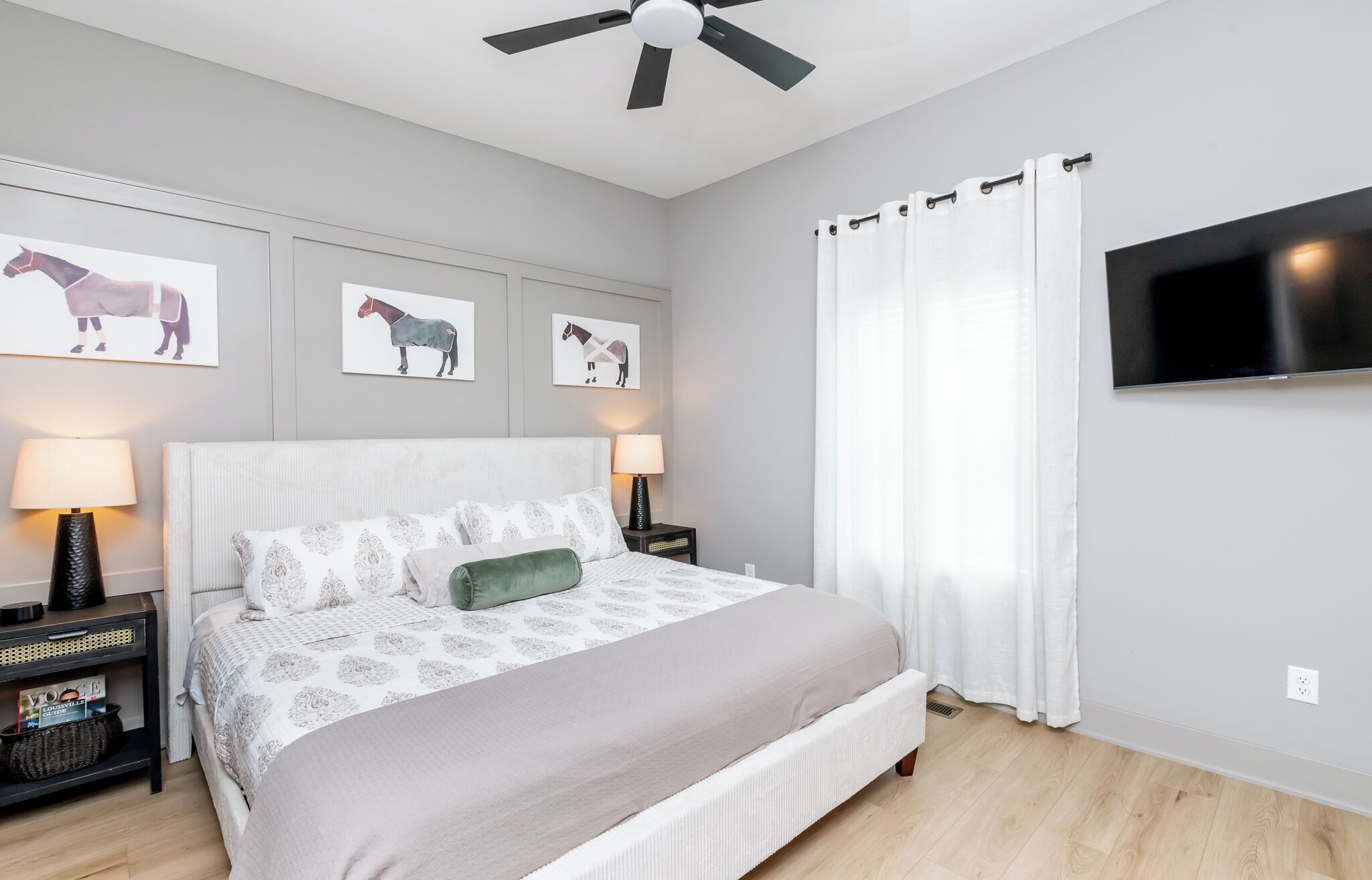 Une chambre à coucher comprenant un lit, deux tables de chevet avec des lampes, un ventilateur de plafond, un téléviseur fixé au mur et des œuvres d’art murales sur le thème des chevaux.