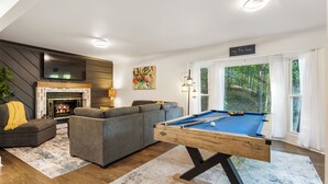 A modern living room with a pool table, a fireplace, a sofa, and a large window offering a view of the outdoors.