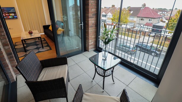 A balcony with a glass door, a wicker chair, a small table with a vase, and a view of a residential street.