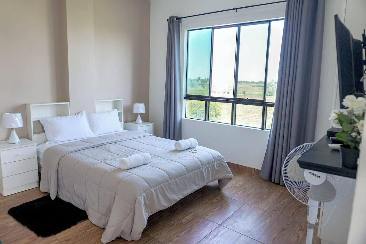 A bedroom with a large bed, white nightstands, a bedside table with a lamp, a window with curtains, and a standing fan.