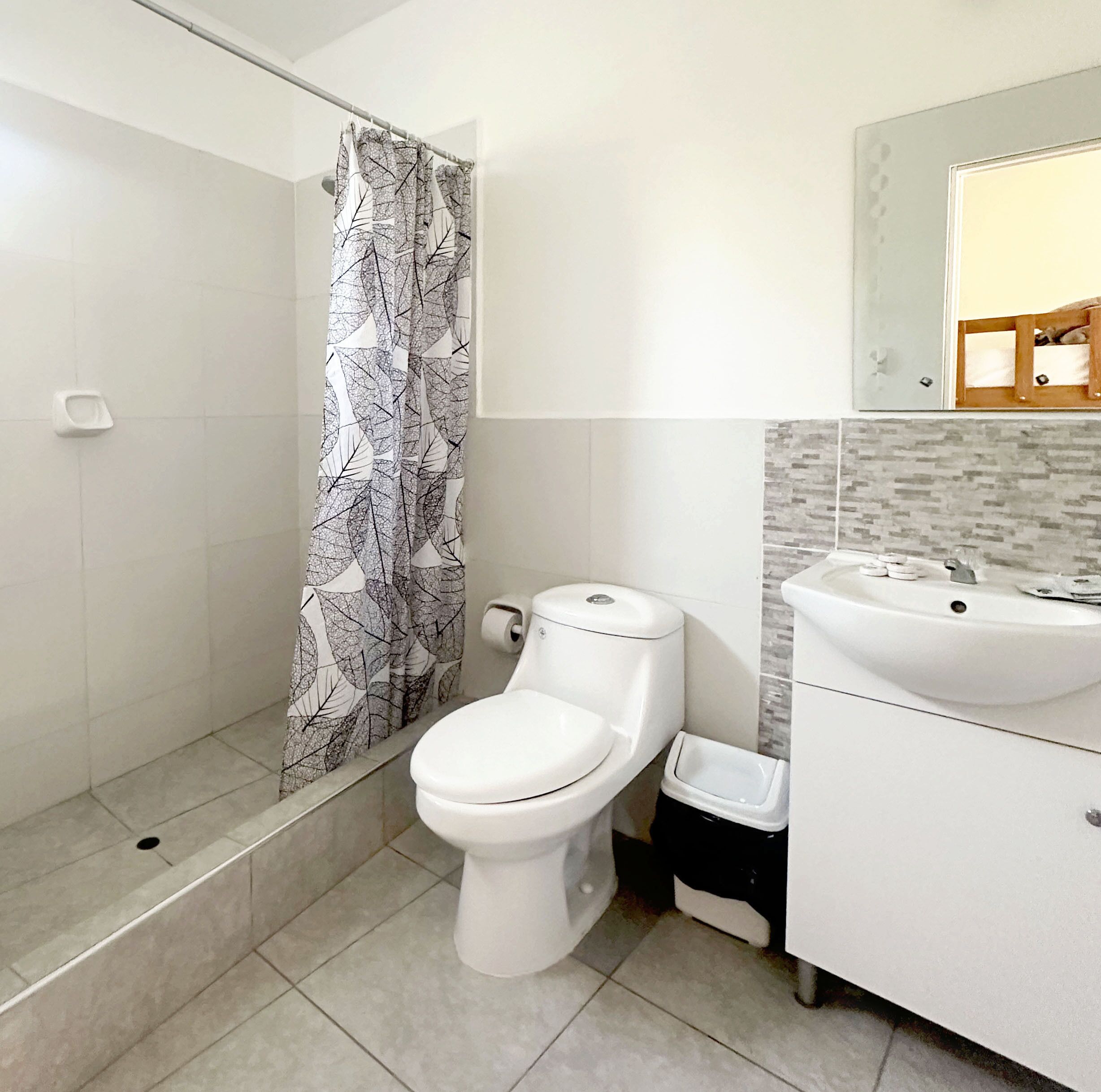 Ein Badezimmer mit Toilette, einer Dusche mit einem Duschvorhang mit Blattmuster, einem Waschbecken und einer gefliesten Wand.