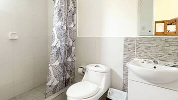 Ein Badezimmer mit Toilette, einer Dusche mit einem Duschvorhang mit Blattmuster, einem Waschbecken und einer gefliesten Wand.
