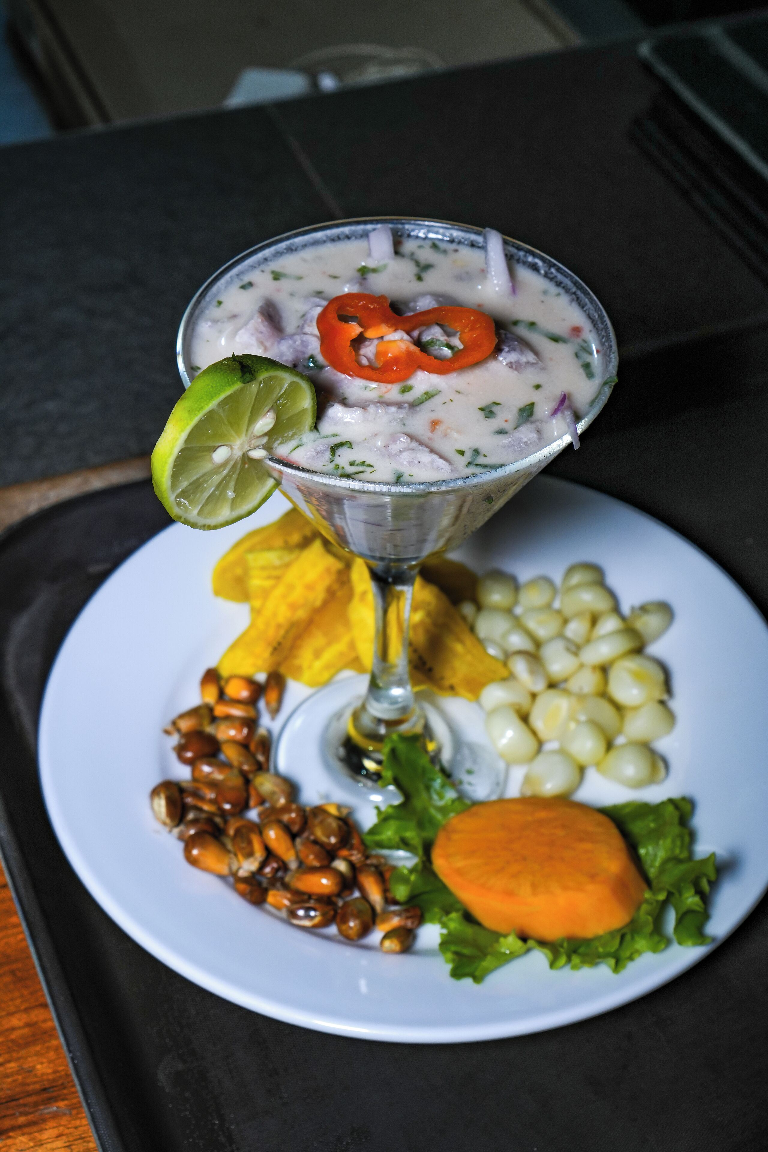 A martini glass with a creamy drink, garnished with a lime wedge and a slice of red pepper, accompanied by a plate with corn, peanuts, a carrot, and a tortilla chip.