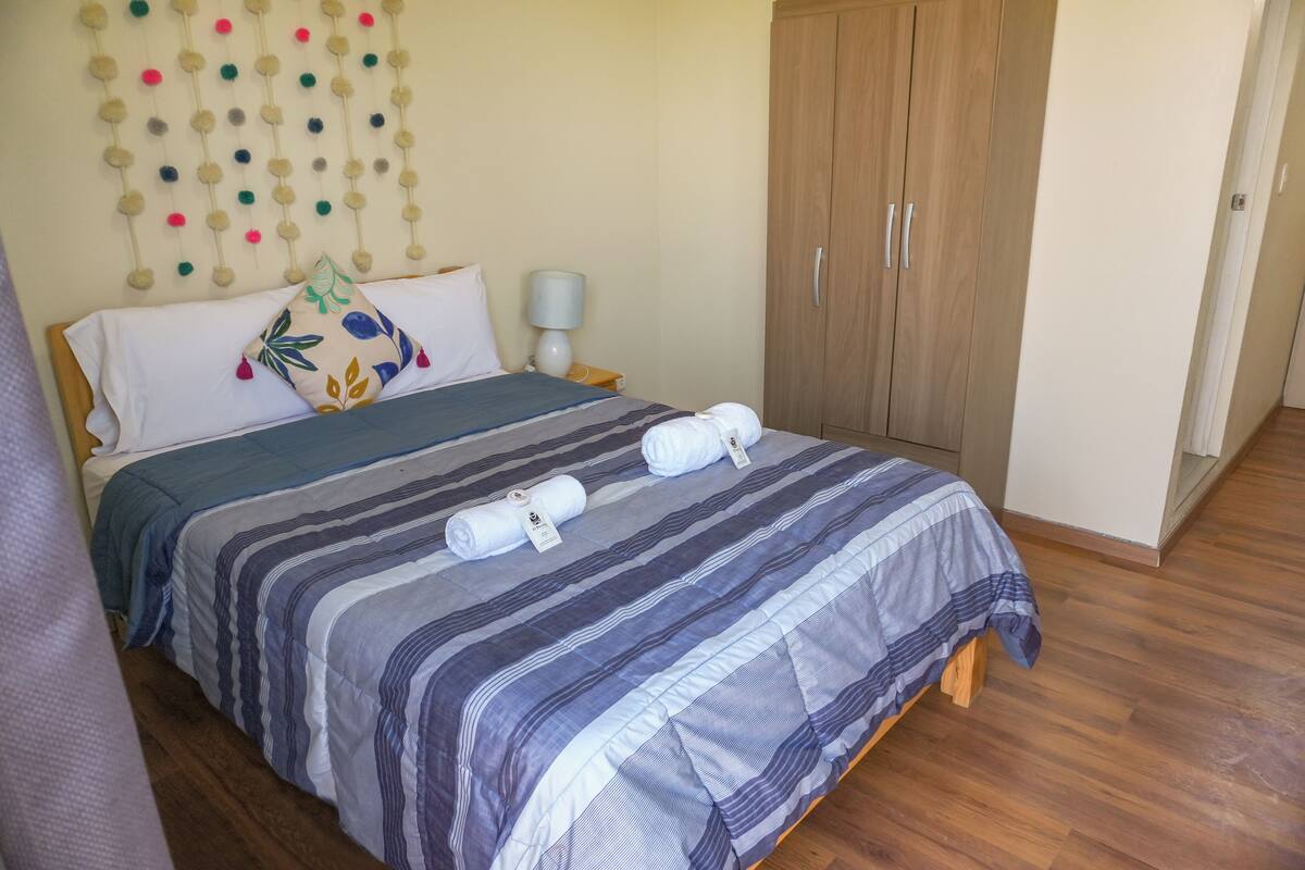 A neatly made bed with striped bedding and two rolled towels. A wall-mounted decorative item hangs above the bed.