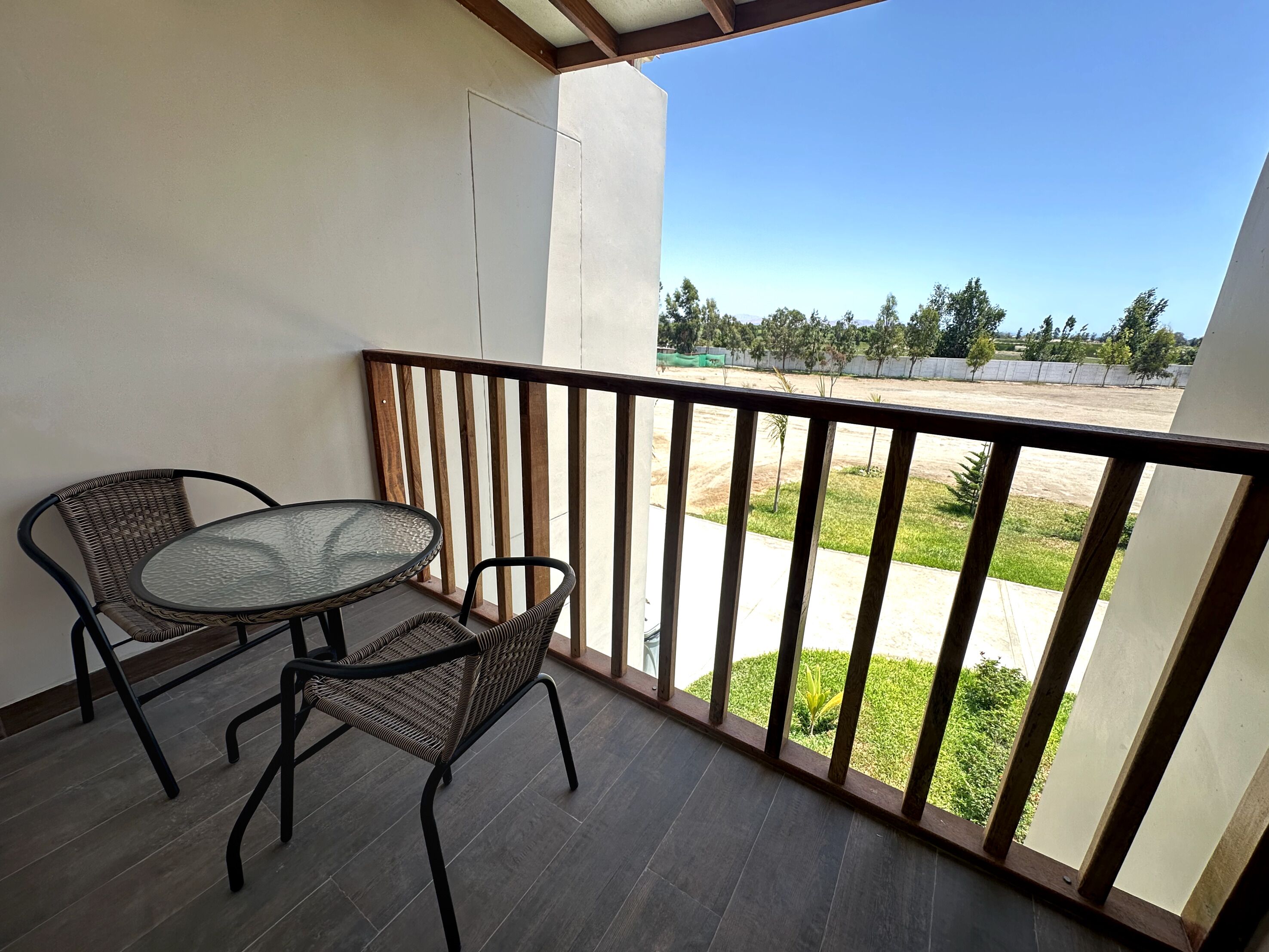 A balcony with a glass table and two wicker chairs, a wooden railing, and a view of a landscaped area with trees and a fence.