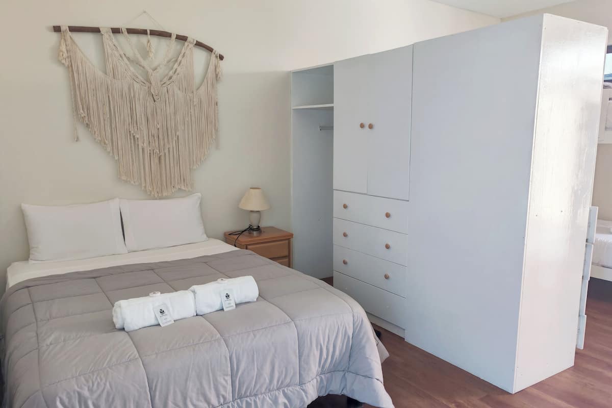 A neatly made bed with a grey duvet, a bedside lamp, and a macrame wall hanging.