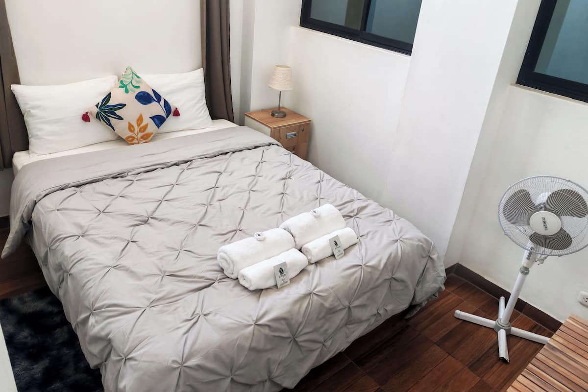 A neatly made bed with a grey quilt, two rolled towels, and a decorative pillow. A small wooden nightstand with a lamp is beside the bed. A standing fan is near the wall.