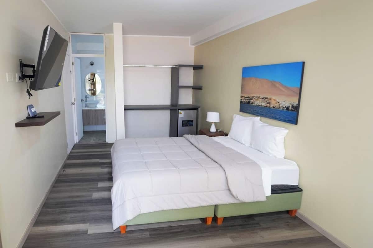 A hotel room with a bed, a TV mounted on the wall, a painting of a landscape, and a small table with a lamp.