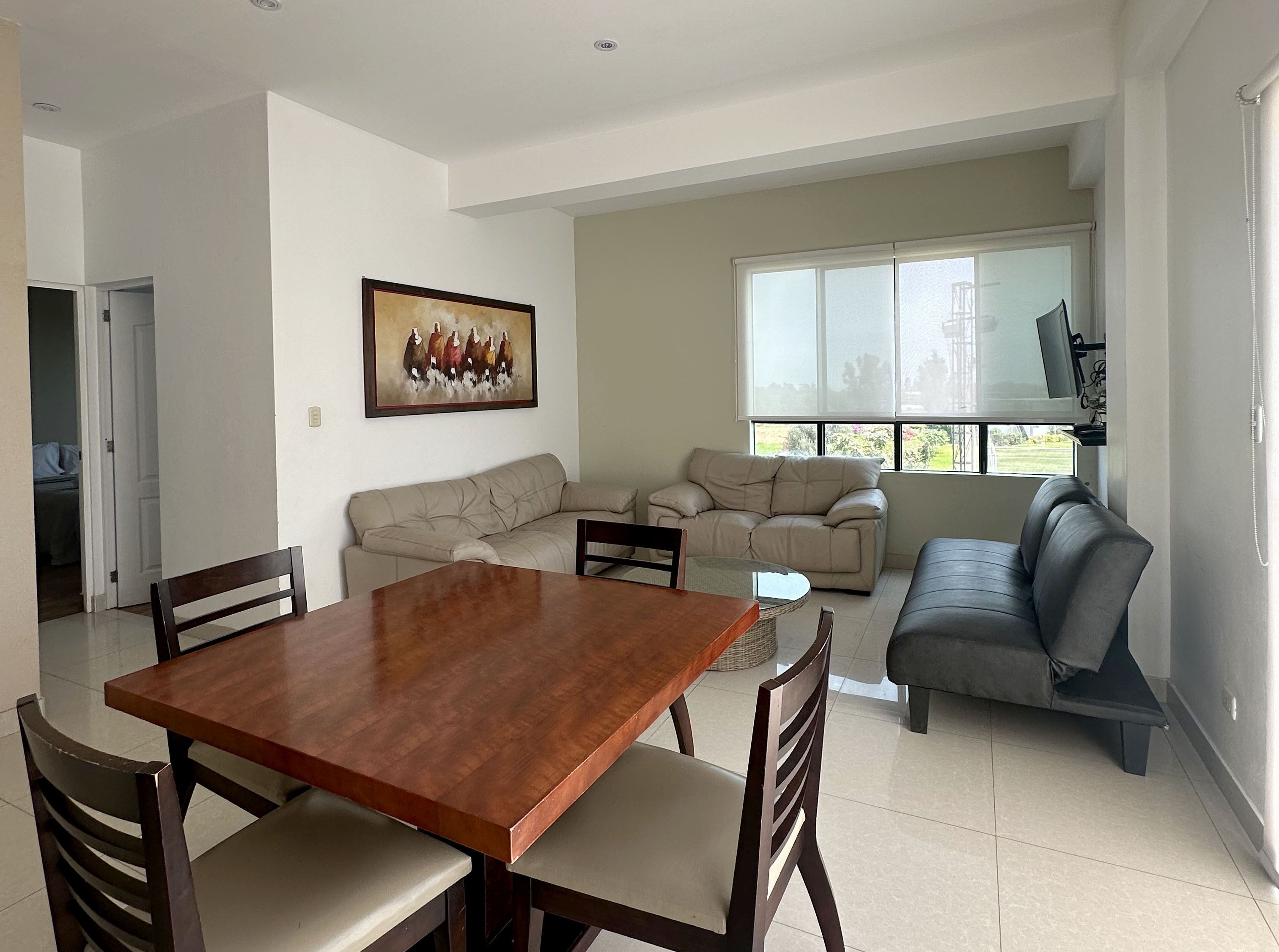 A dining area with a wooden table and chairs, a beige sofa, and a gray sofa. There is a painting on the wall and a window with blinds.