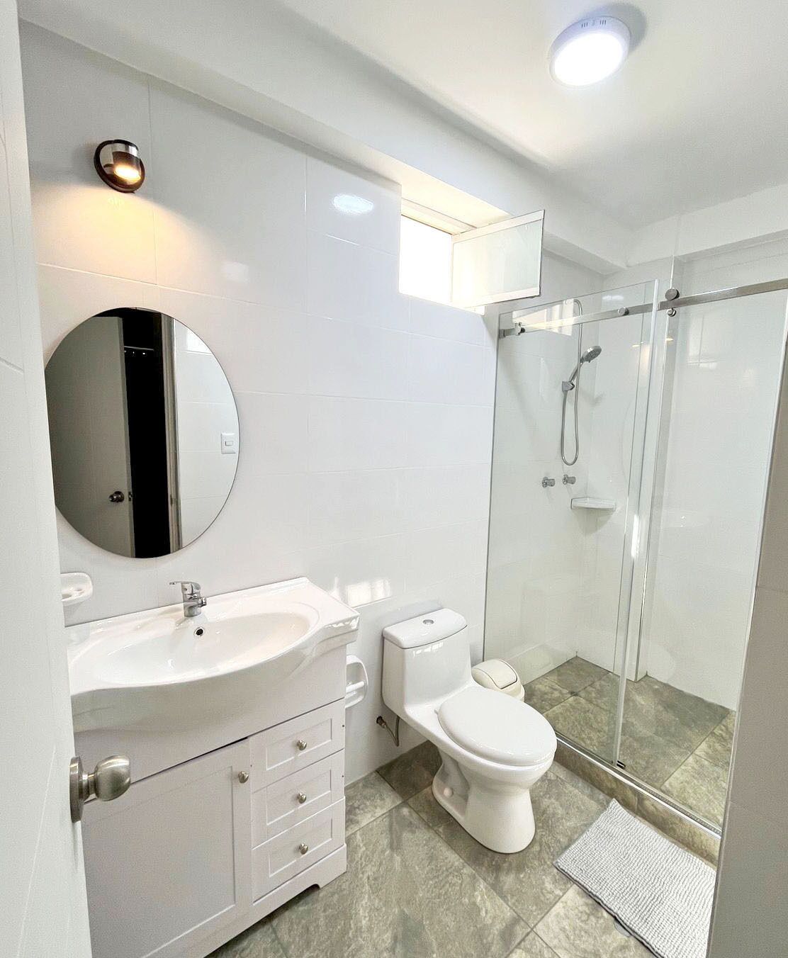 A modern bathroom with a round mirror, a white sink, a toilet, and a glass-enclosed shower.