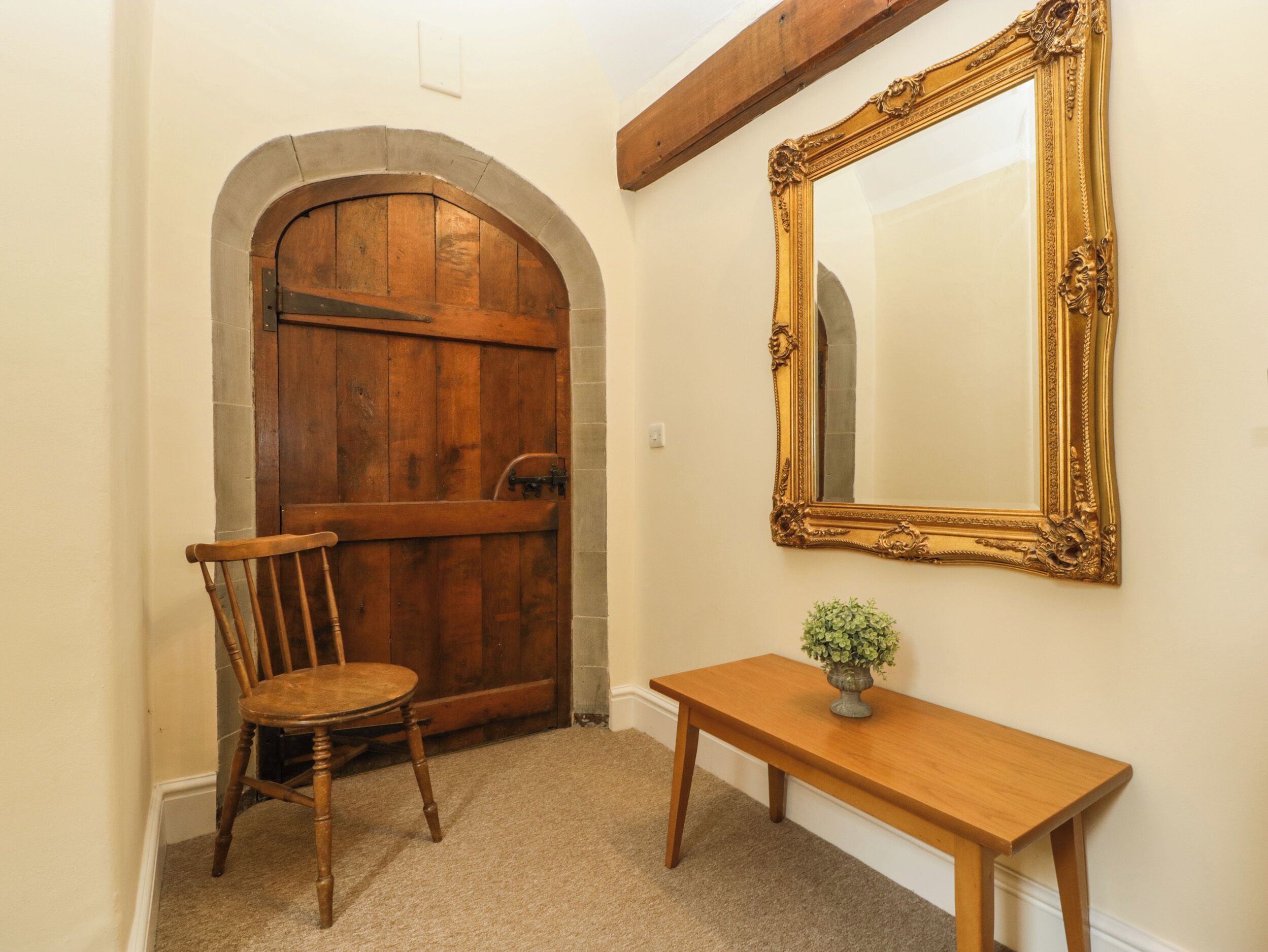 A wooden door with an arched top, a wooden chair, a wooden table with a vase, and a large ornate mirror on the wall.