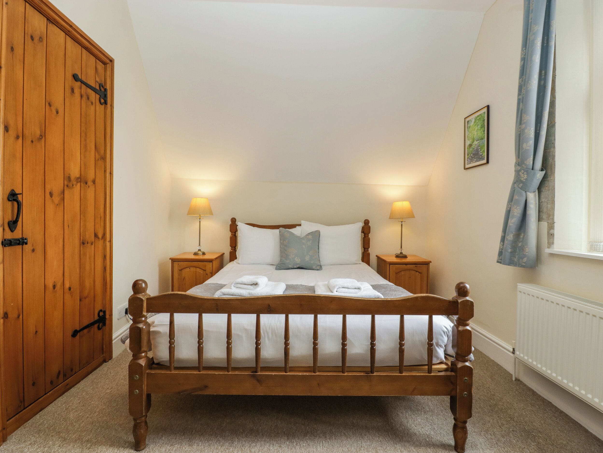 A bedroom with a wooden four-poster bed, two bedside tables with lamps, a wooden wardrobe, and a window with curtains.