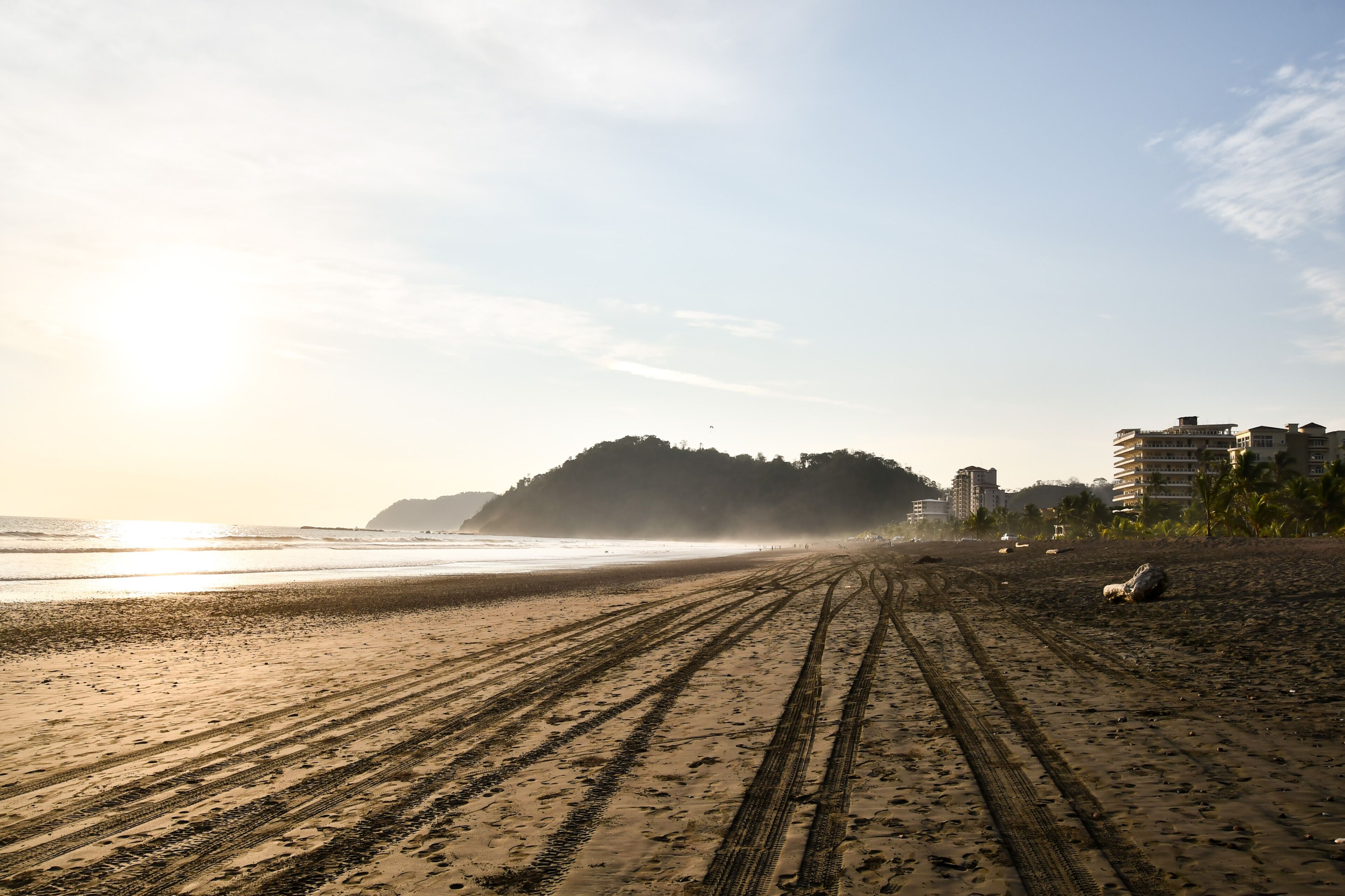 A beach with tire tracks leading to the water, a distant hill, and buildings in the background.