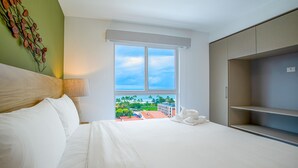 Une chambre d’hôtel avec un lit, une fenêtre donnant sur des palmiers et des immeubles, une armoire et un élément décoratif mural.