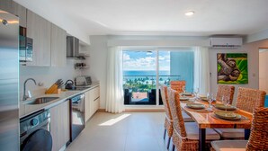 Une cuisine moderne équipée d’un lave-linge intégré, un coin repas avec des chaises en osier et un balcon offrant une vue sur l’océan.