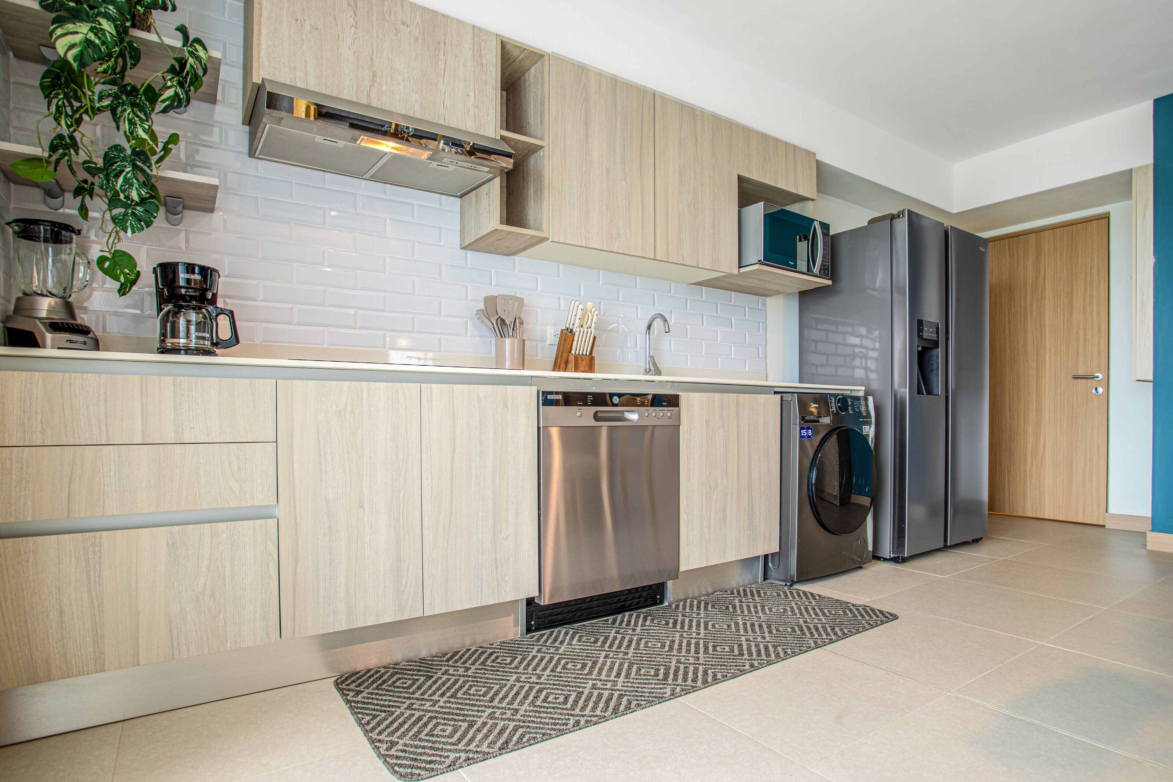 Una cocina moderna con armarios de madera clara, lavavajillas de acero inoxidable, lavadora integrada y un refrigerador grande.